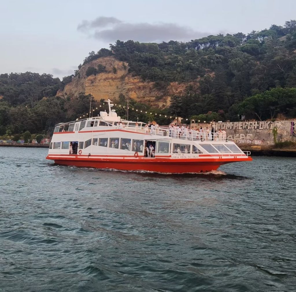LVT Lisbon event boat cruising on the Tagus river at dusk below green cliffs – Barco de eventos LVT Lisbon a navegar no rio Tejo ao entardecer